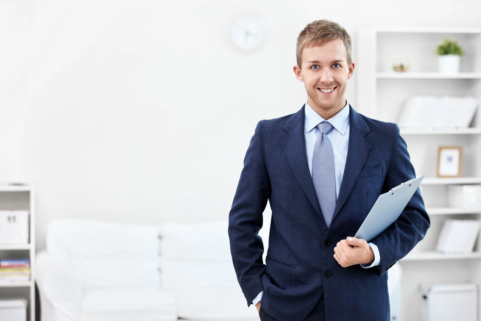 A smiling man in a suit holds a clipboard in a bright office